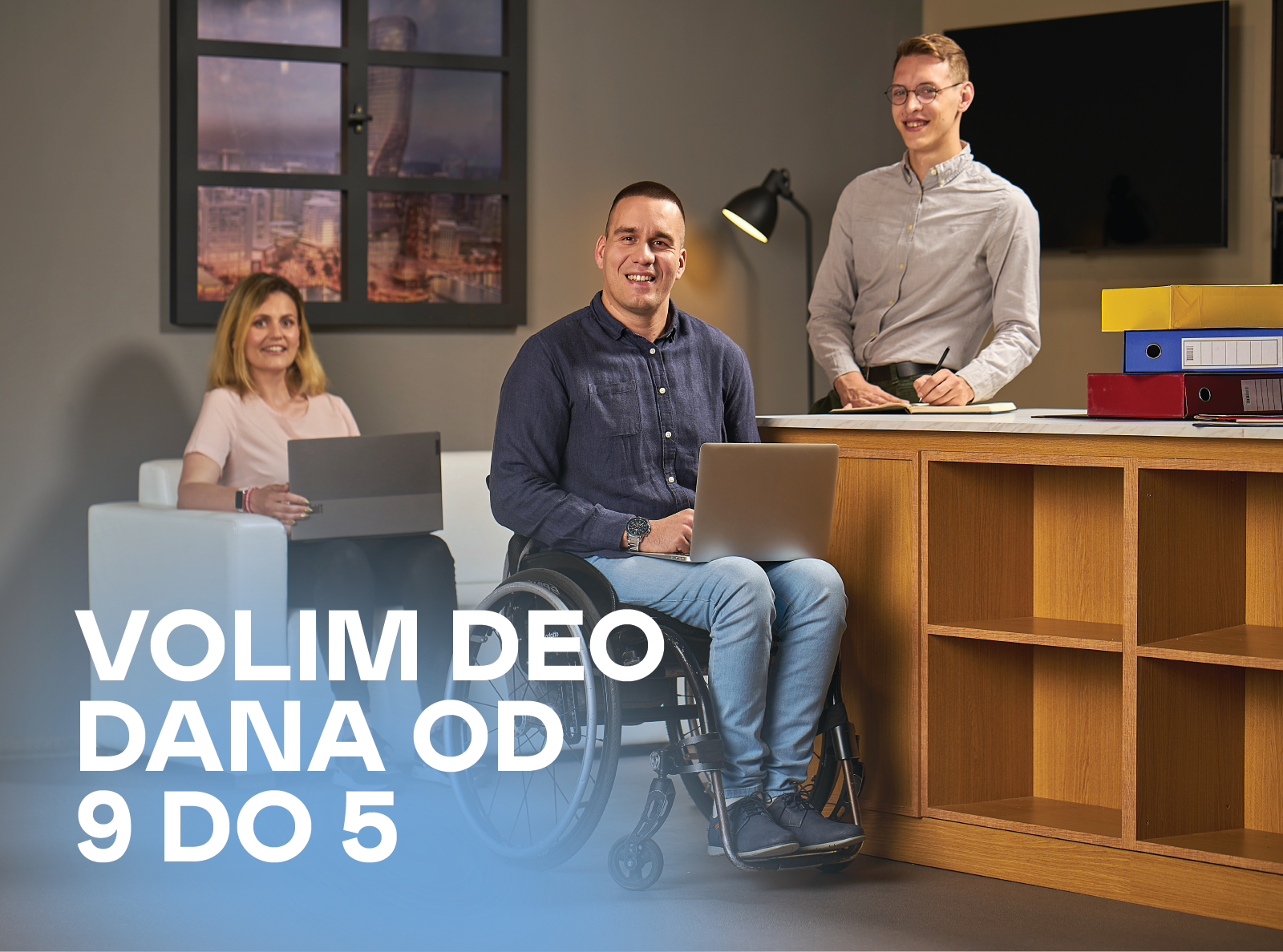  U radnom okruženju muškarac korisnik kolica sedi ispred radnog stola sa laptopom u krilu i osmehuje se. Pored njega stoji mladić za radnim stolom i nešto zapisuje, dok se iza nalazi žena koja sedi na beloj fotelji sa laptopom u krilu. Preko slike stoji tekst: „Volim deo dana od 9 do 5.