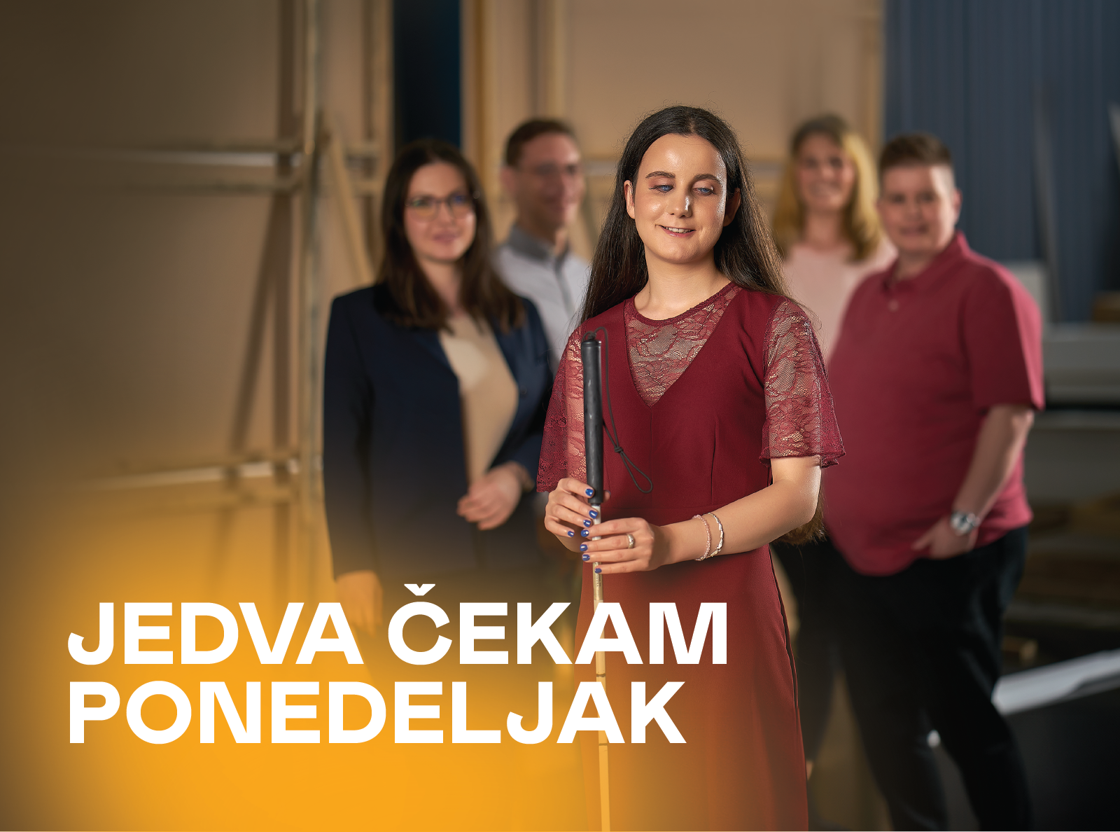  Mlada žena u bordo haljini stoji nasmejana u prvom planu i drži beli štap za orijentaciju namenjen osobama sa oštećenjem vida. Iza nje se nalaze kolege, tri žene i jedan muškarac, u poslovnom okruženju. Na slici piše: „Jedva čekam ponedeljak.“