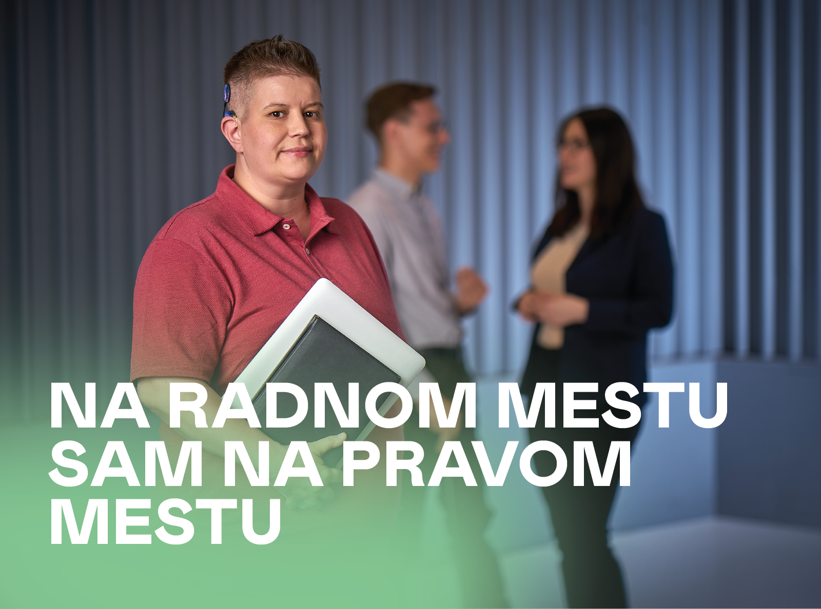  Ženska osoba kratke kose u crvenoj polo majici, sa kohlearnim implantom iza desnog uha, drži fasciklu i blago se osmehuje. Iza nje, u prijatnoj i opuštenoj atmosferi radnog prostora, nalaze se kolege – muškarci i žene. Preko slike stoji tekst: „Na radnom mestu sam na pravom mestu.”